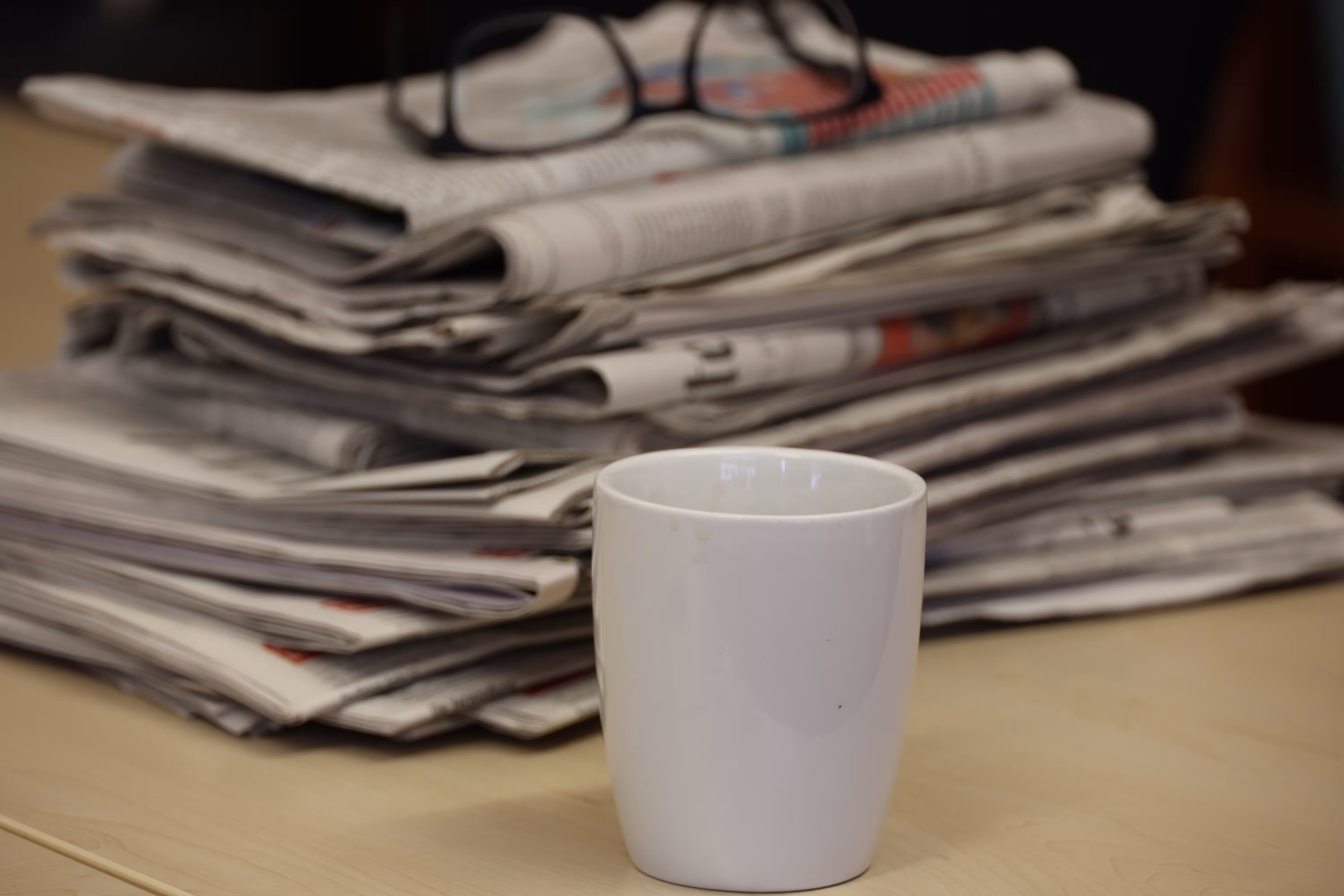 Stack of newspapers with glasses set aside. A white cup stands in front of it.