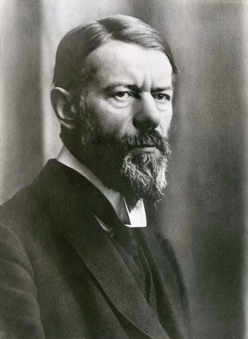 Black-and-white photo of a man with a long full beard in three-quarter profile. He has a side parting and is wearing a suit. It is Max Weber.