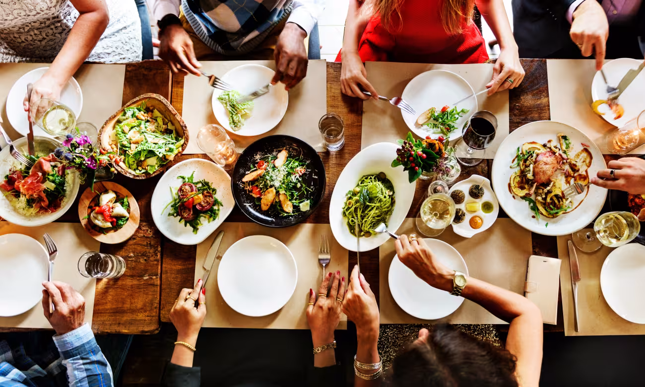 A table set for dinner, seen from a bird's eye view. In the center are dishes from different cultural cuisines. Cut off at the edges and recognizable only by their arms, the diners are in the process of placing various dishes on their plates.