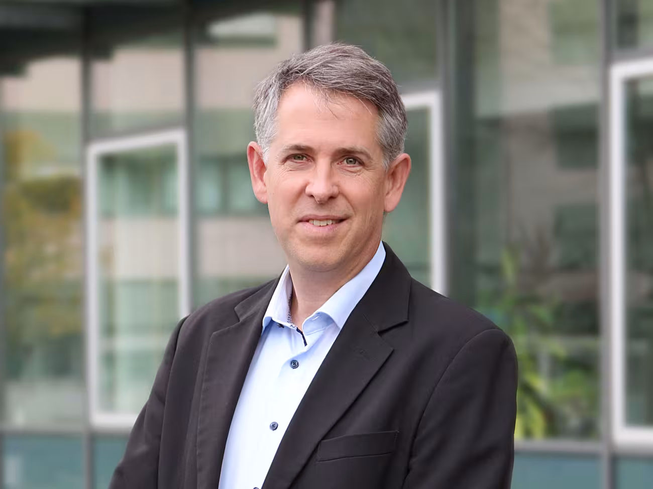 Stefan Pfister, a man with short gray hair combed to the side, smiles at the camera. He is wearing a light-colored shirt and a dark blazer over it. 